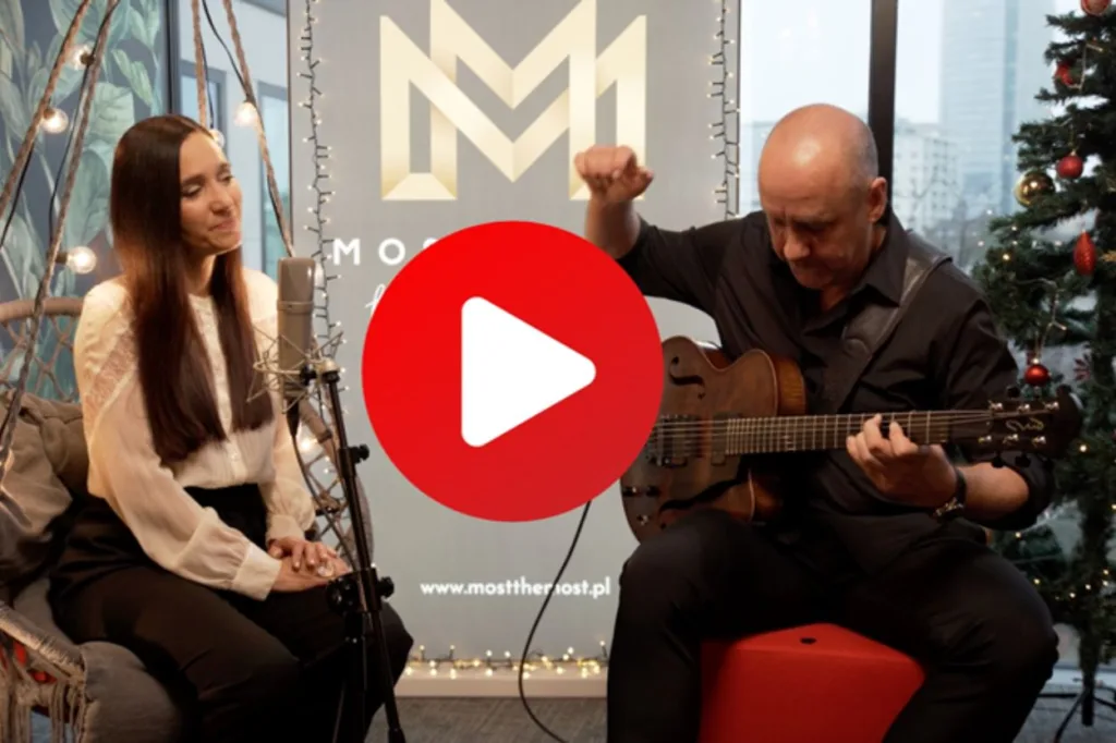 Na zdjęciu kadr z nagrania wideo kolędy. Dorota Miśkiewicz siedzi na podwieszonym fotelu. Obok Marek Napiórkowski gra na gitarze. W centralnej części przycisk "play".