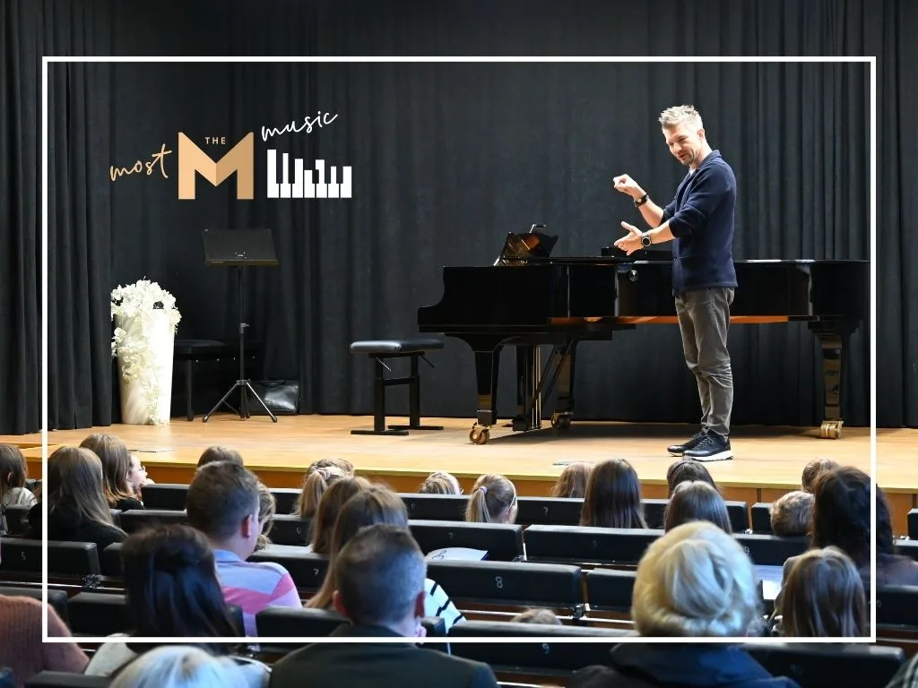 Na zdjęciu warsztaty Most the Music w Rudzie Śląskiej. Na scenie Adam Sztaba opowiada i gestykuluje. Na widowni dzieci i młodzież chórów Most the Music oraz ich rodzice i opiekunowie.