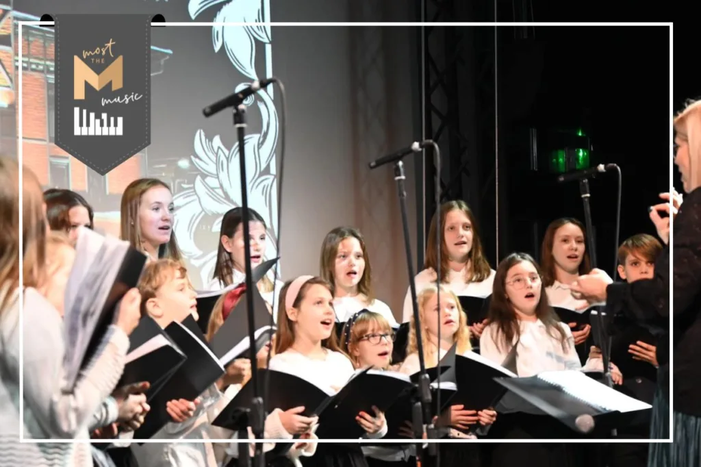 Na zdjęciu dzieci i młodzież z chóru Most the Music z Rudy Śląskiej w trakcie występu. Ubrani na galowo, w rękach trzymają zeszyty z nutami. W lewym górnym rogu logo programu Most the Music.