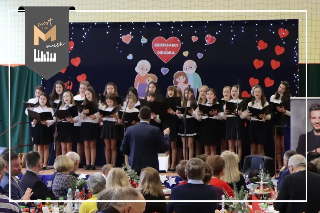 Występ chóru Most the Music z Binarowej/Libuszy. Na scenie ubrane na galowo dzieci i młodzież. W tle granatowa dekoracja z postaciami babci i dziadka, miedzy nimi czerwone serce. Przed samą sceną chórmistrz. W lewym górnym rogu logo programu Most the Music.