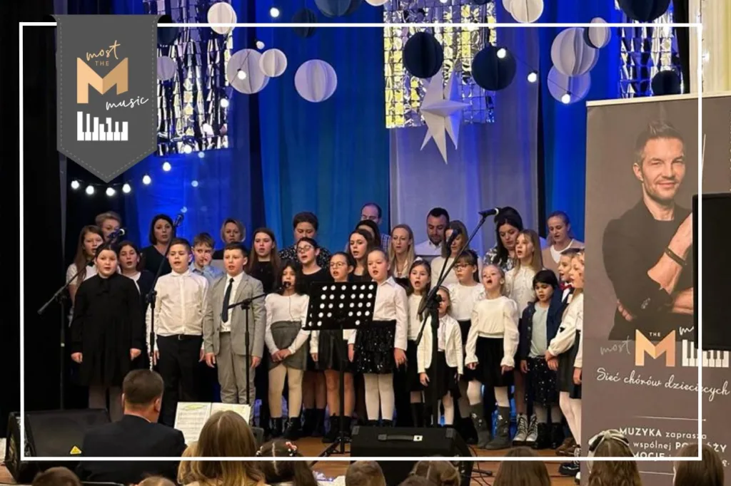 Występ chóru Most the Music z Cieszanowa. Na scenie ubrane na galowo dzieci i młodzież z gościnnym udziałem rodziców. W tle granatowa dekoracja z połyskującymi srebrnymi elementami (wiszące kółka, gwiazdy). Przed samą sceną siedzi chórmistrz, który gra na instrumencie klawiszowym. w Prawej części zdjęcia widoczny jest baner programu Most the Music, na którym widać zdjęcie Adama Sztaby, dyrektora artystycznego programu. W lewym górnym rogu logo Most the Music.