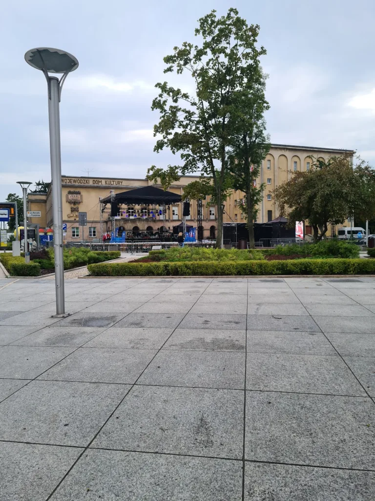 Fragment ulicy miejskiej wraz ze skwerem, drzewami oraz budynkiem Domu Kultury.