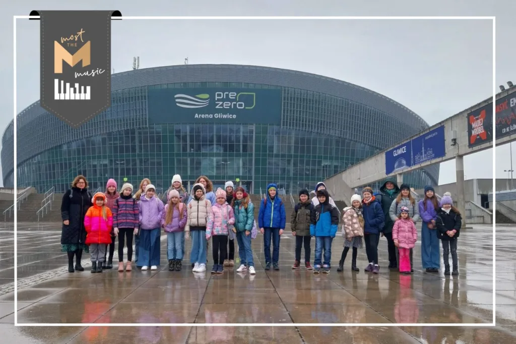 Zdjęcie grupowe chóru Most the Music przed PreZero Arena Gliwice. Deszczowa pogoda. W lewym górnym rogu logo programu Most the Music.