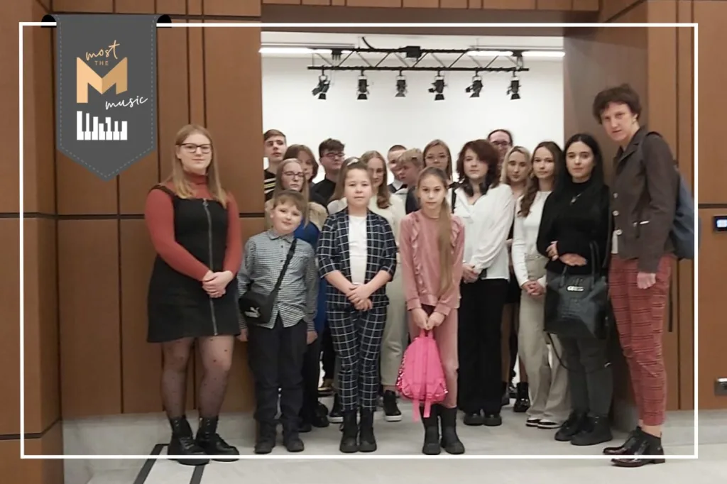 Zdjęcie grupowe wewnątrz budynku filharmonii. Na zdjęciu dzieci i młodzież w towarzystwie opiekunów. W lewym górnym rogu logo programu Most the Music.