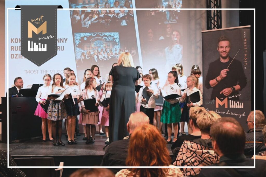Dzieci śpiewające na scenie. Przed nimi stoi chórmistrzyni i kieruje śpiewem. Po lewej stronie gra mężczyzna na instrumencie klawiszowym. W tle ozdobna grafika. w lewym górnym rogu logo programu most the music.