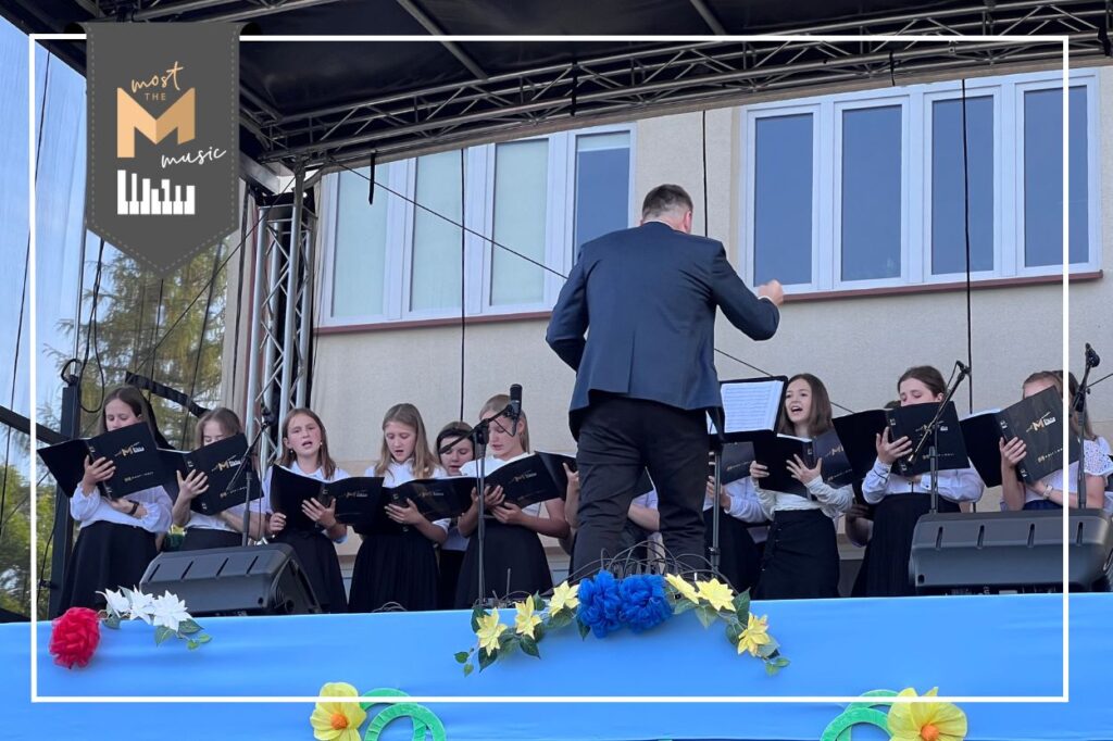 Młodzież na scenie podczas występu. Przed nimi dyrygent. Logo most the music.