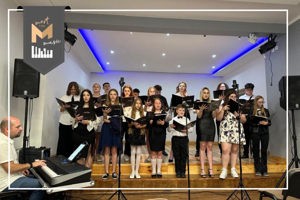 Dziecięco-młodzieżowy chór podczas występu na scenie. Po lewej stronie mężczyzna gra na instrumencie klawiszowym. W lewym górnym rogu logo programu most the music.
