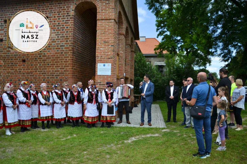 Zdjęcie z otwarcia murowańca. Po lewej stronie lokalny zespół ludowy, na środku przemawia wójt gminy, po prawej grupa ludzi - lokalna społeczność. W tle zabytkowy murowaniec.